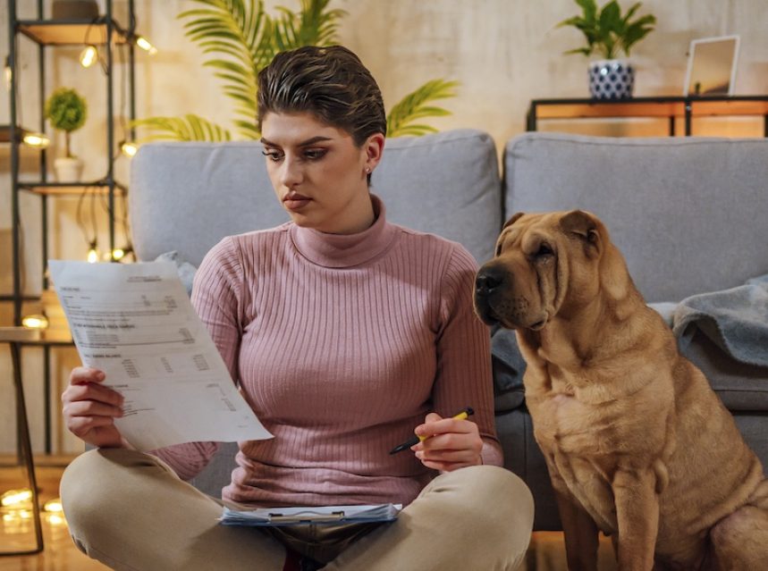 Young woman sits on the floor with her dog, looking over bills.