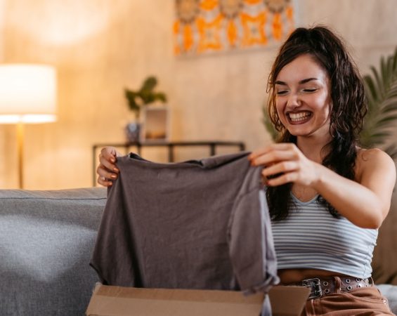 A smiling woman pulling a new shirt out of a package while sitting on a couch.