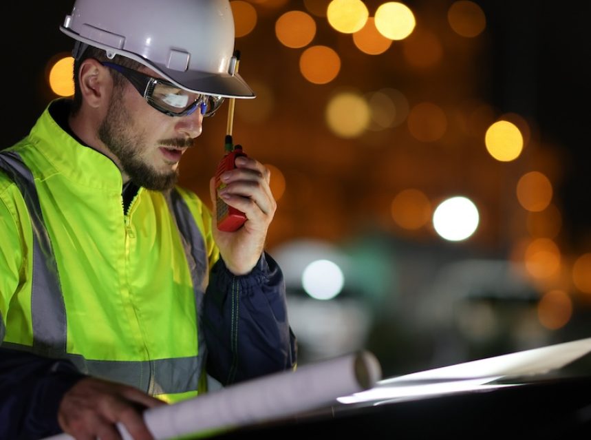 An electrical engineer wearing a reflective jacket and hard hat is illuminated against a dark background while using a walkie talkie.